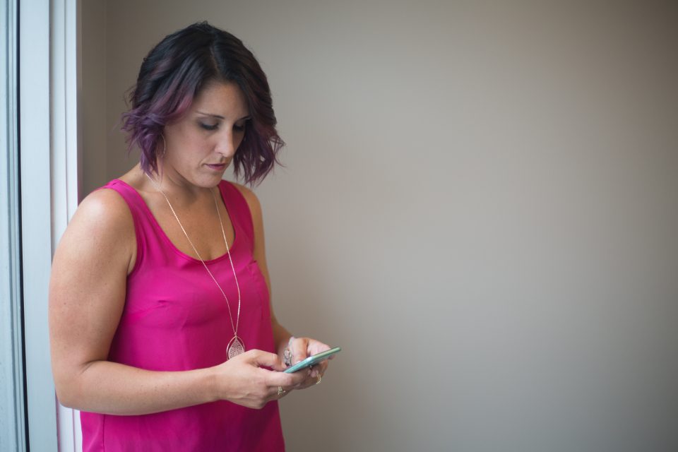 Mulher de blusa rosa digitando no celular, encostada numa parede
