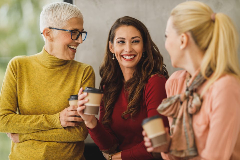 Três colegas de trabalho conversando e sorrindo, com copos de café nas mãos