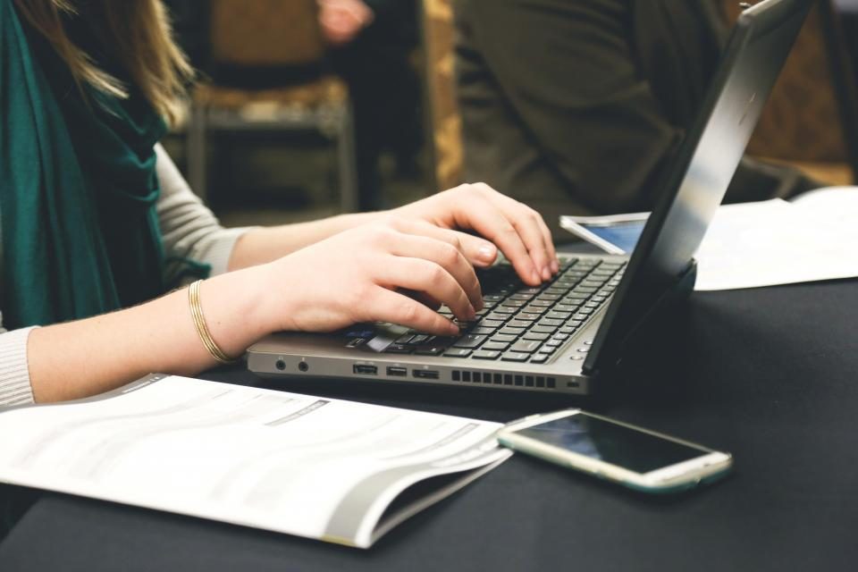 Mãos digitando num notebook, com um caderno aberto e um celular ao lado