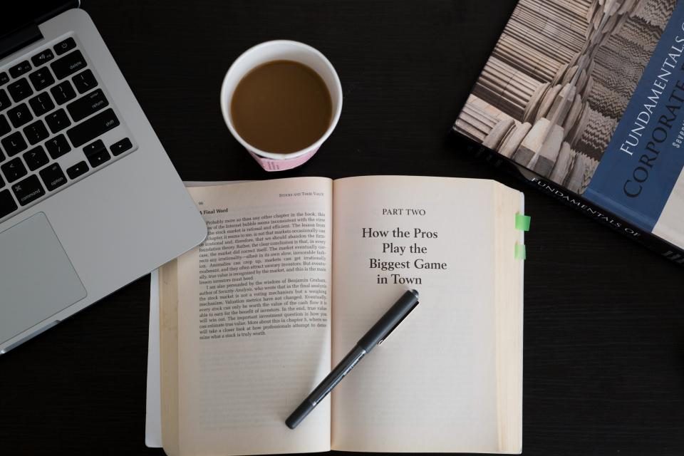 Livro aberto sobre uma mesa escura, ao lado de um notebook, uma caneta e uma xícara de café