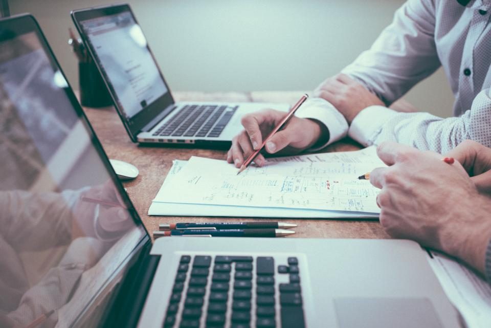 Mãos fazendo anotações num caderno ao lado de dois notebooks numa mesa de trabalho